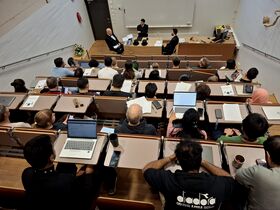 Students in a tiered lecture hall watch three speakers at the front, many using laptops and taking notes.