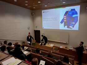 Lecture hall audience watching a thesis defence on heated textiles, with presenters in formal black suits.