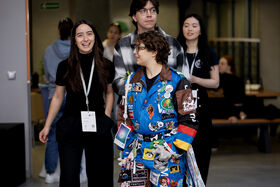 Group of people indoors. One wearing a blue jumpsuit covered with various patches and badges, others dressed casually.