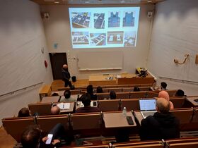 Lecture hall with students watching a man present photos of equipment on a large screen