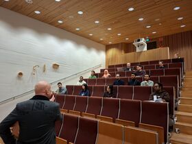 Lecturer addresses small, diverse audience in tiered wood-panelled lecture hall with red seats.