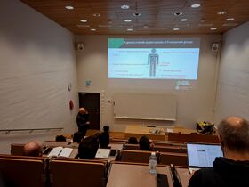Lecture hall with students watching a slide on e-textile systems; lecturer stands by the front whiteboard.