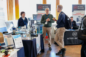 Exhibition booth with various equipment on tables and banners. Three people are having a conversation and one is seated with a laptop.
