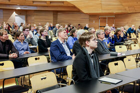 People sitting and listening attentively in a wooden panelled auditorium. Front tables have notebooks and pens.