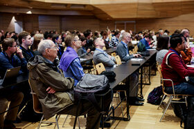 A large room filled with people seated at tables, attentively listening. Wooden walls and high ceiling are visible.