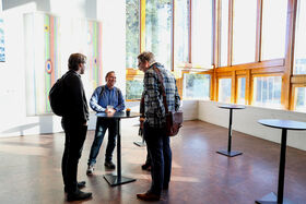 Three people stand at a tall table in a bright room with large windows and abstract artwork on the wall.