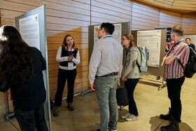 People are discussing research posters in a conference setting with wooden walls. Some are wearing badges and lanyards.