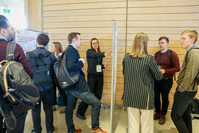 Group of people discussing around a poster in a wooden-panelled room. Several attendees have backpacks and conference badges.