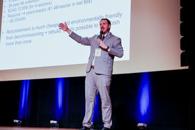 A man in a grey suit is giving a presentation on stage with a microphone. A slideshow is projected behind him.