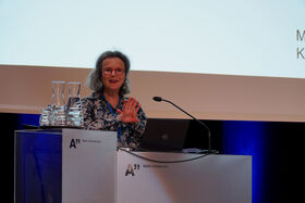 A person speaking at a podium with a laptop and water jugs, under a screen at Aalto University.