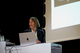 A person at a podium with an Apple laptop, presenting at Aalto University. A glass of water is on the desk.