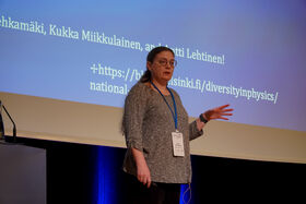 Person presenting on stage with a large screen showing text and URLs behind them.