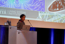Person at a podium with a laptop, speaking. Background shows a screen with the words 'Living Systems Institute'.
