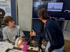 Nainen sinisessä puserossa osoittaa ruudulle istuvalle miehelle. Pöydällä on teknisiä laitteita.