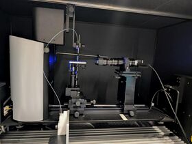 Laboratoriolaitteisto, jossa on optisia ja mekaanisia osia mustalla alustalla. Näkyvissä on johtoja ja sininen valo.