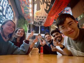 Kaveriporukka ravintolapöydässä tekee sormisydämiä värikkäiden kattoverkkojen alla