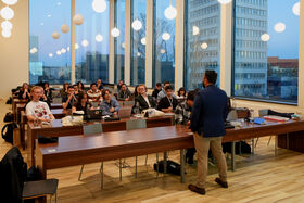 A man is presenting to a seated group in a modern classroom. Large windows show an urban scene. Pendant lights hang from the ceiling.