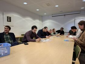 People sit around a light wooden table with plastic bags, papers, and cards. A blue basket is on the table.