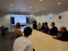 A woman presents in a conference room with six attendees seated around a table. A presentation slide is visible on a screen.
