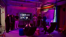People give a presentation on a large screen in a purple-lit modern room with seated listeners.
