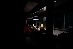 Dim office with two people working at bright screens under a black hanging lamp at night