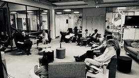 Group of people in a modern office lounge, seated on sofas in a semicircle, listening to a speaker.