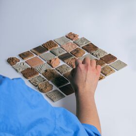 Person in blue sleeve arranging small textured brown and grey tiles on a white surface