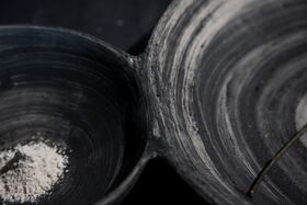 Close-up of two connected dark ceramic bowls, one with white powder, the other with a thin stick.