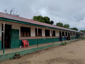 Long single-storey school building with green doors, sandy yard and a few people standing outside.