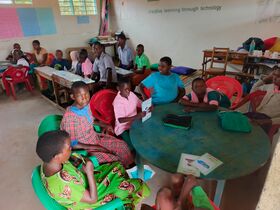 Children sit around tables in a classroom with red and green chairs, reading and talking together.