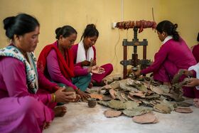 Women in pink clothes sit on the floor, stacking dried leaves beside a hand press against a yellow wall