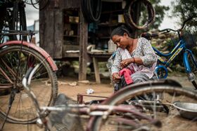 Person repairing a bicycle on a dusty ground outside a wooden shed, surrounded by old bikes and tools