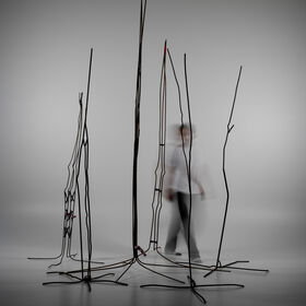 Blurred person walks among tall, thin metal rod sculptures in a grey studio space