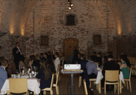 People seated at tables in a brick-vaulted hall listen to a speaker standing at the left.