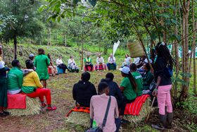 Outdoor group meeting in a forest clearing, people sit in a circle on hay bales with bright blankets.