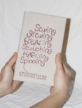 Hands holding a beige textile craft book with red wavy title text on a white fabric background