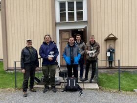 Viisi henkilöä droneineen seisoo puisen rakennuksen edessä, lähettyvillä pieni sininen patsas
