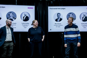 Three people stand in front of two large judge-introduction screens at an indoor event.