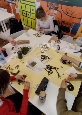 People at a workshop table crafting with green seaweed-like material on yellow sheets