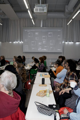 People sit at white tables in a classroom, watching a projection on a grey screen, drinks and snacks on desks