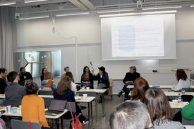 Seminar room with people seated at tables, listening to a panel in front of a large projected slide