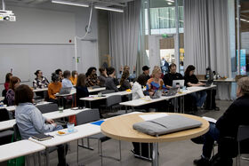 People in a modern classroom sit at long tables with laptops, watching a speaker out of frame.