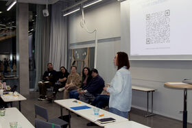 Person in blue shirt speaking to seated group in modern classroom with QR code projected on screen
