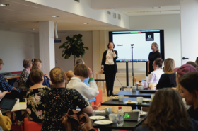 People at a seminar watch two speakers by a large screen reading “Kiitos!” in a bright meeting room