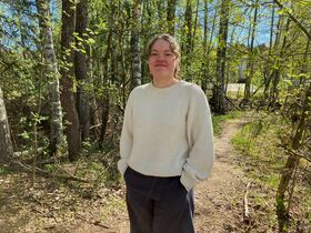 Person in a light jumper and dark trousers stands on a forest path in spring sunlight.