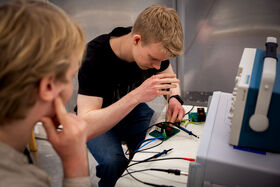 Two students work with cables and circuit boards beside a large blue and grey lab device
