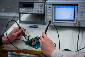 Two people test an electronic circuit board with probes, next to grey lab instruments and a blue graph screen.