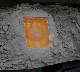 Translucent orange face mould set in wet resin, surrounded by grey sand or dust.