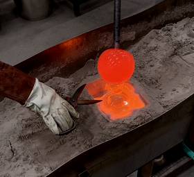 Gloved worker shaping bright orange molten glass sphere in a sand mould with metal shears