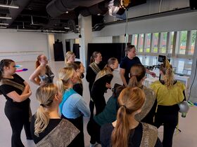 Group of dancers in glittering costumes watch instructions on a screen in a bright studio